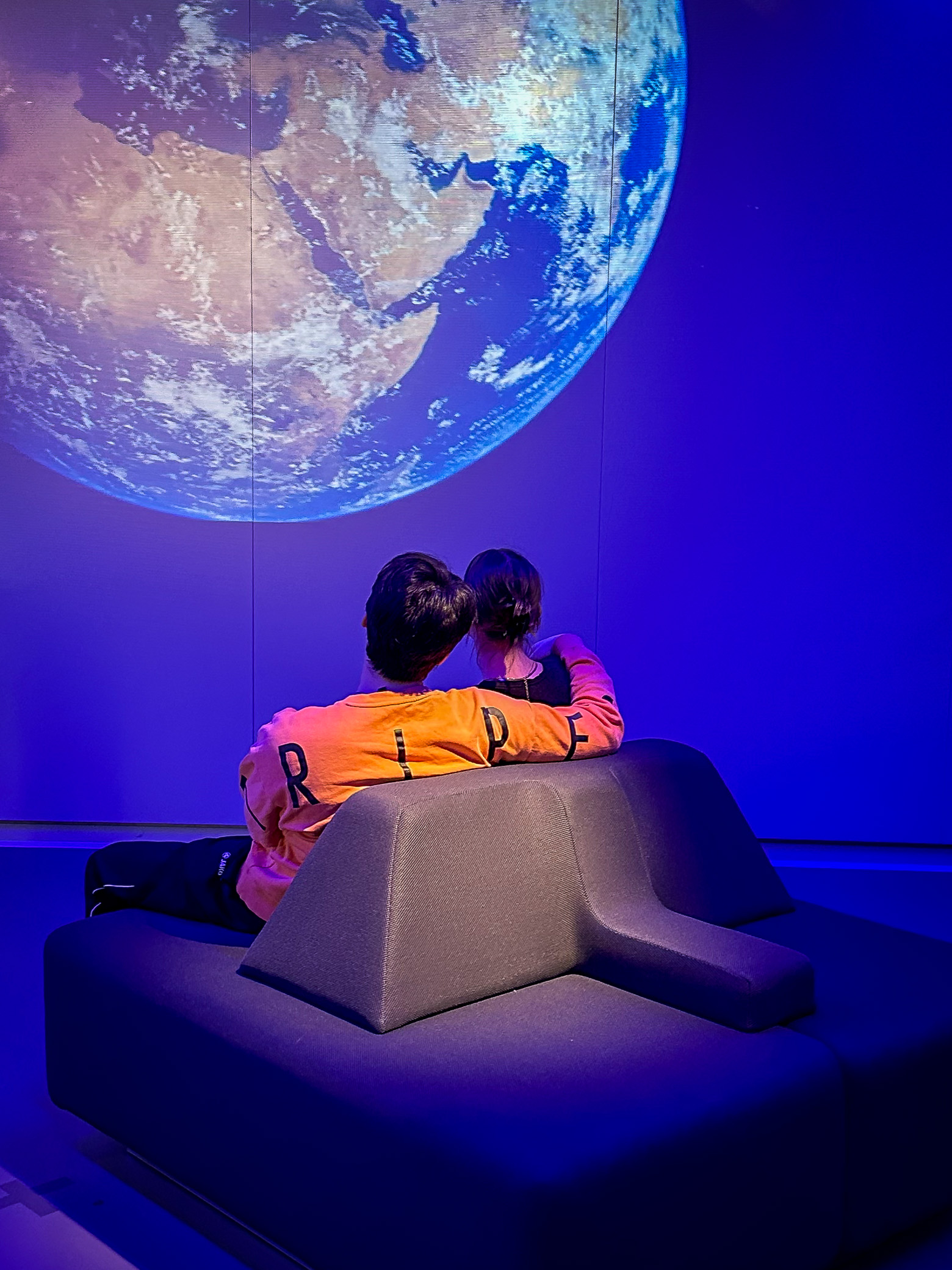 A young couple sit in a lounge chair casually observing a projection of planet earth - Casual Observations