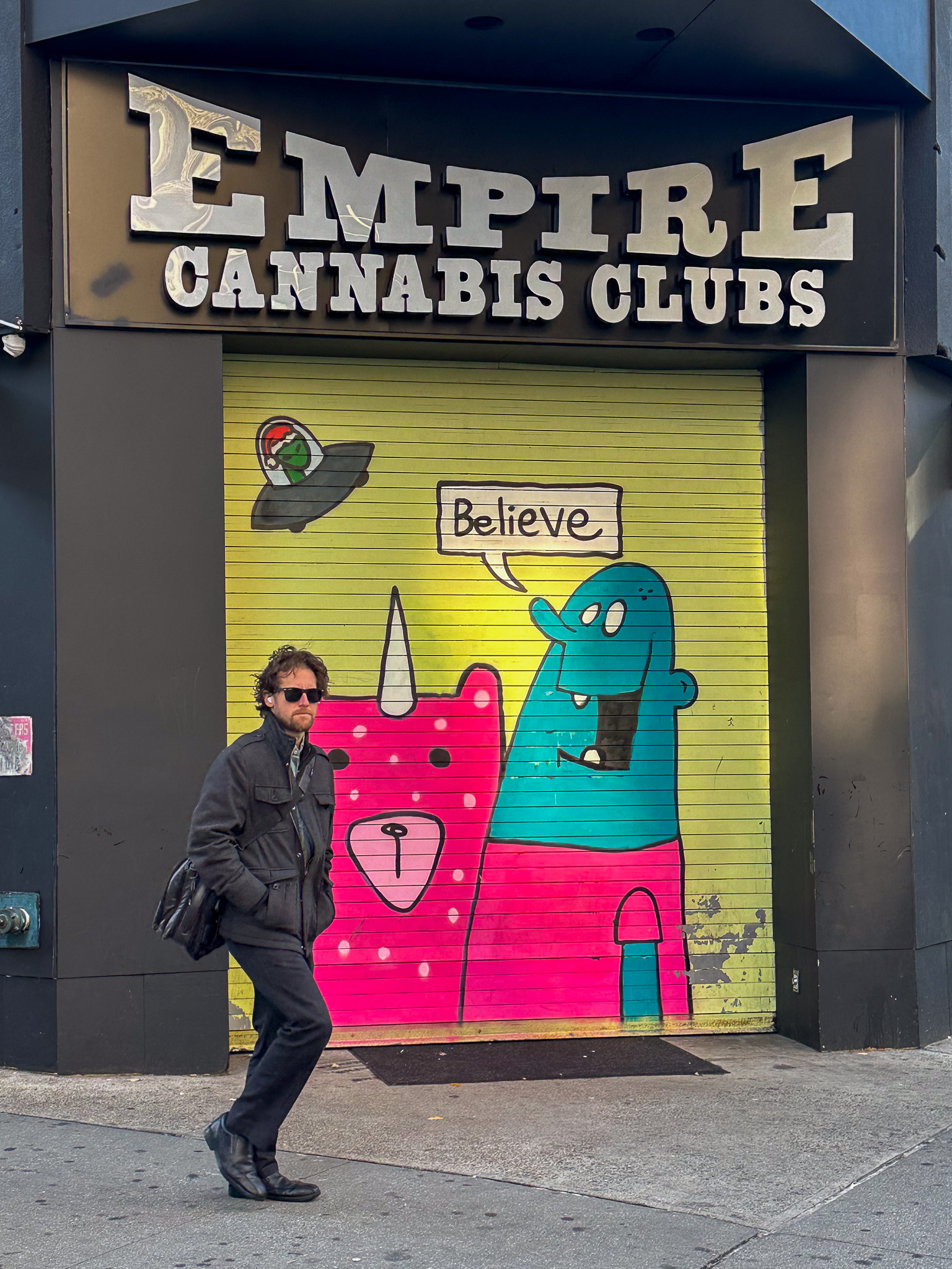 A man walking in front of a cannabis club with a cartoon on the door and an alien saying believe
