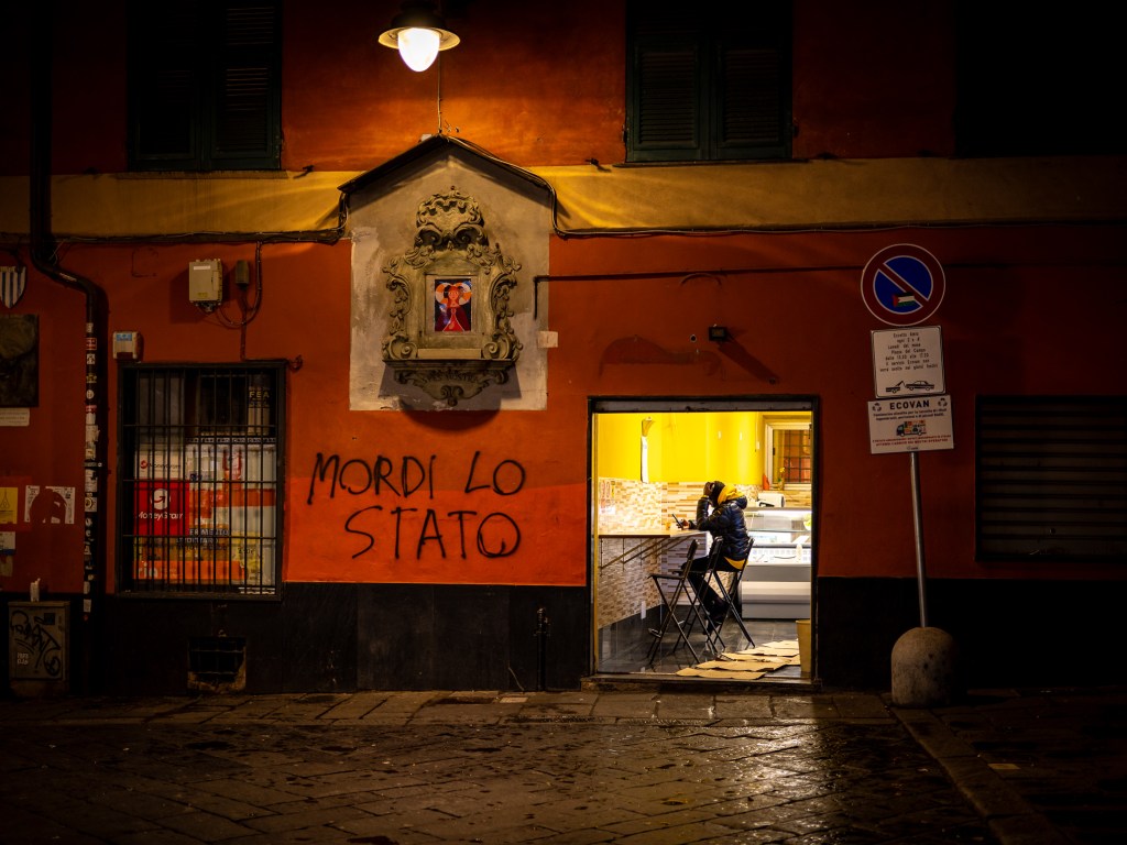 Man sitting in a lit shop looking into his cellphone next to a graffiti that translates to bite the state