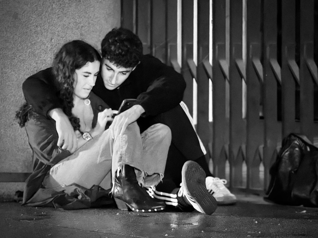A young couple sitting on a doorstep, oblivious to the world, silently sharing a view in their cellphone