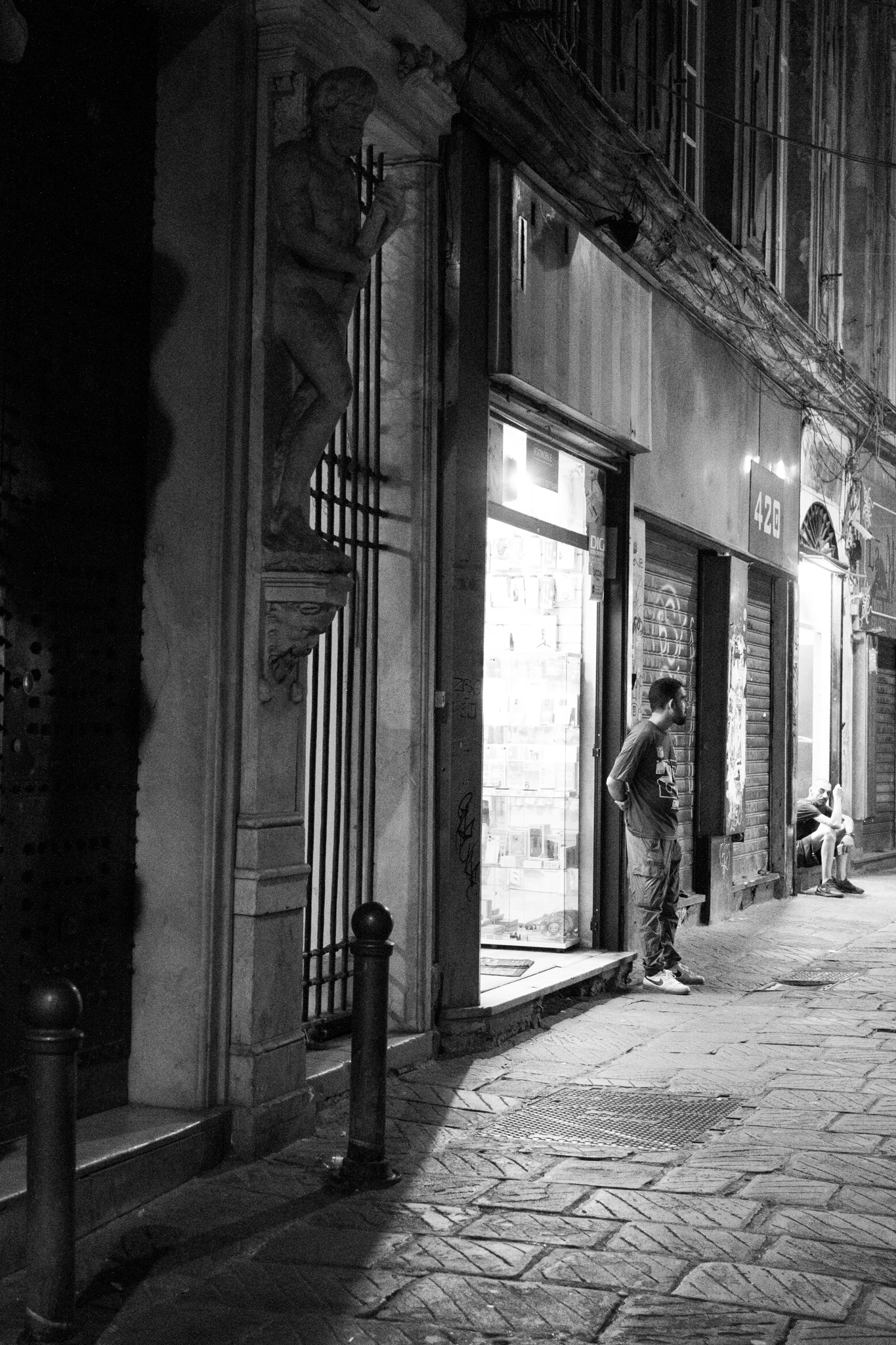 A statue on a house entry, a standing and a sitting man silently observe the pedestrians in an alley in Genoas Centro Storico